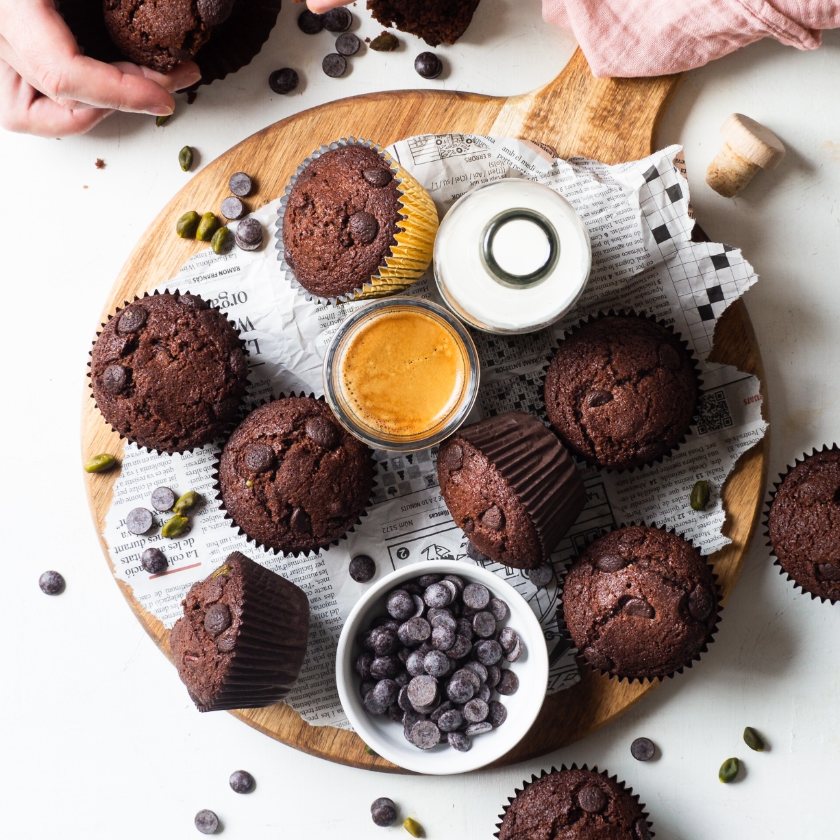 Chocolate chip muffins on a board with scattered chocolate chips and seeds, an espresso and some milk
