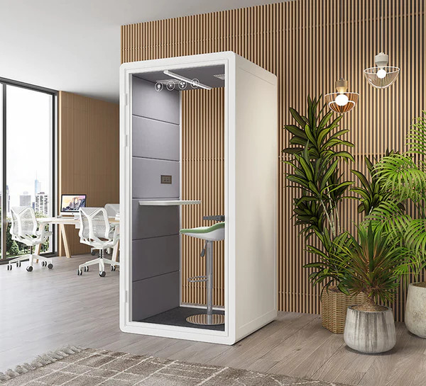 A sleek white privacy booth positioned in a modern office space with wooden vertical accents. The booth's interior reveals a high-standing desk, overhead lighting, and a gray acoustic wall panel. In the background, an open workspace with multiple desks and chairs is visible, along with large windows showcasing city views. A tall potted plant adds a touch of nature to the scene.
