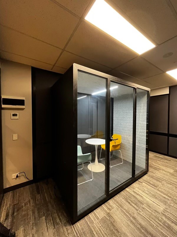 A modern privacy booth set in a dimly lit office area. The booth features dark framing with clear glass walls, showcasing its interior which includes a white circular table and two chairs in contrasting colors of muted teal and bright yellow. The interior wall of the booth is textured in gray, adding a touch of sophistication. Outside, the office design includes wood-patterned flooring, overhead rectangular lights, and dark wall panels, creating a cohesive and contemporary ambiance.