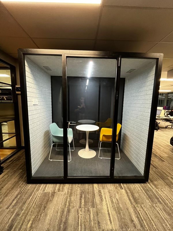 An elegant privacy booth placed in a modern office environment. The booth is framed in dark metal and enclosed with transparent glass, revealing an interior that consists of a round white table flanked by a muted teal and a bright yellow chair. The interior walls are adorned with a light gray brick pattern. The booth's sliding door is slightly ajar, and the broader office space outside is visible, featuring wood-textured flooring and other office furniture in the background. The reflection of the photographer is subtly captured in the glass.