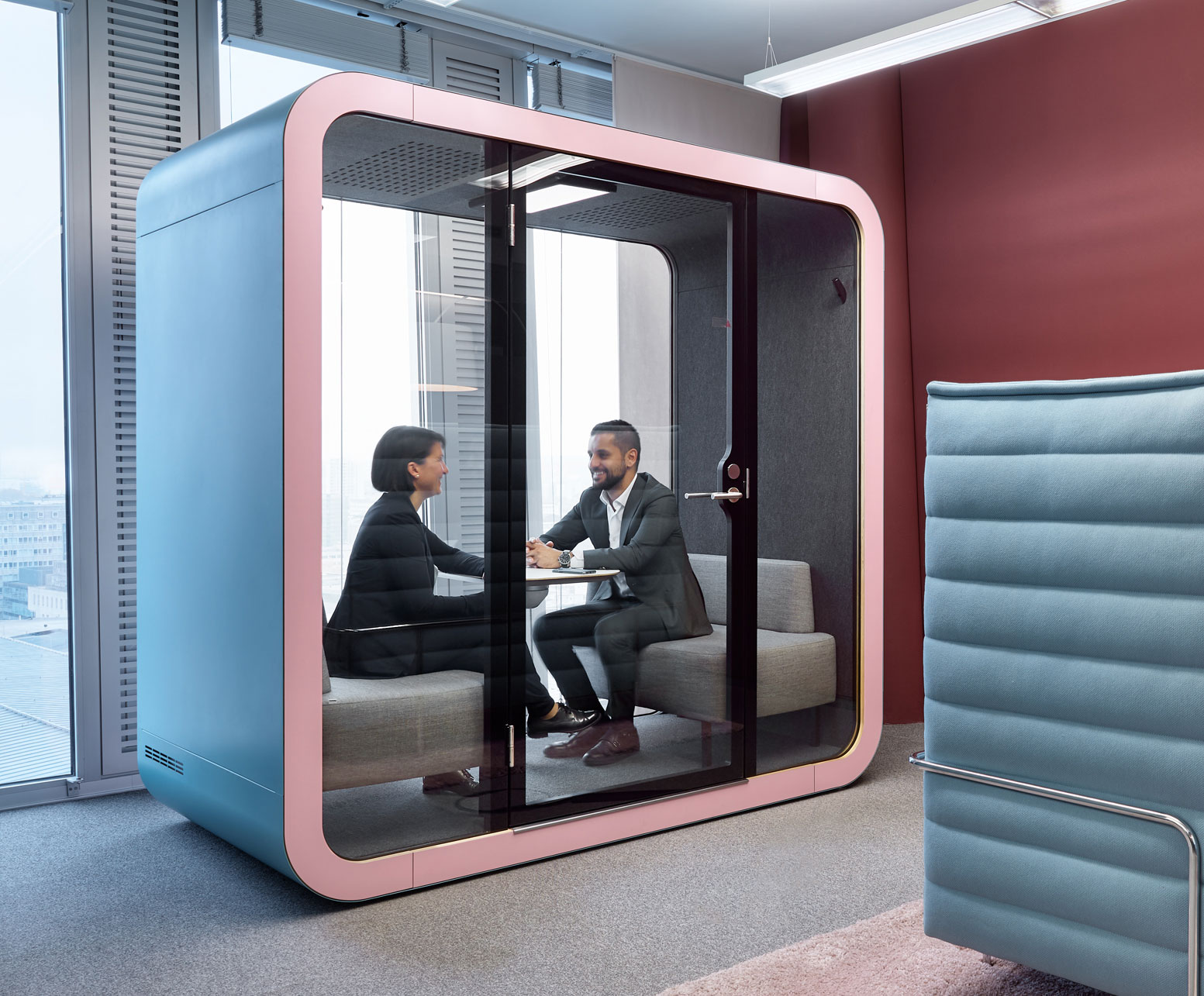 A modern pastel blue privacy booth situated in a stylish office setting. Inside the booth, two professionals are engaged in a focused conversation, seated across a table from each other. The booth's transparent glass walls allow for a clear view of its interior, which is complemented by cushioned seating. Adjacent to the booth is a red wall and a blue padded partition, with large windows on the left offering a glimpse of the cityscape outside.